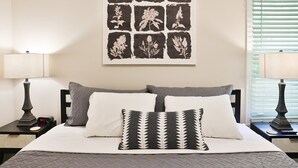Ein ordentlich eingerichtetes Schlafzimmer mit einem Bett, zwei Nachttischen, einem an der Wand befestigten Bild mit floralen Mustern und einem Fenster mit Jalousien.