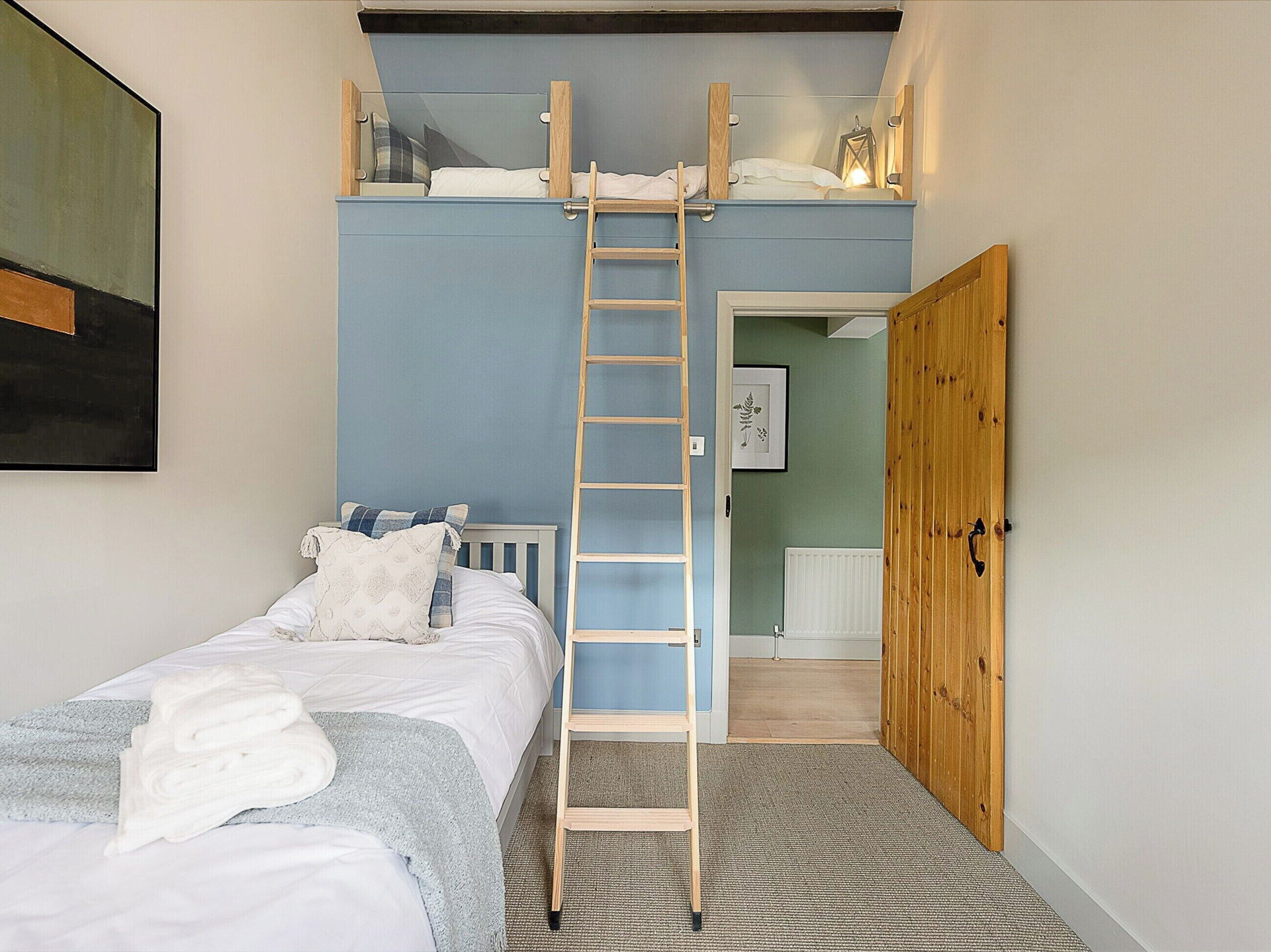 Una camera da letto con un letto a castello, una scala, un letto con lenzuola bianche, una parete blu, una porta in legno e un quadro appeso al muro.