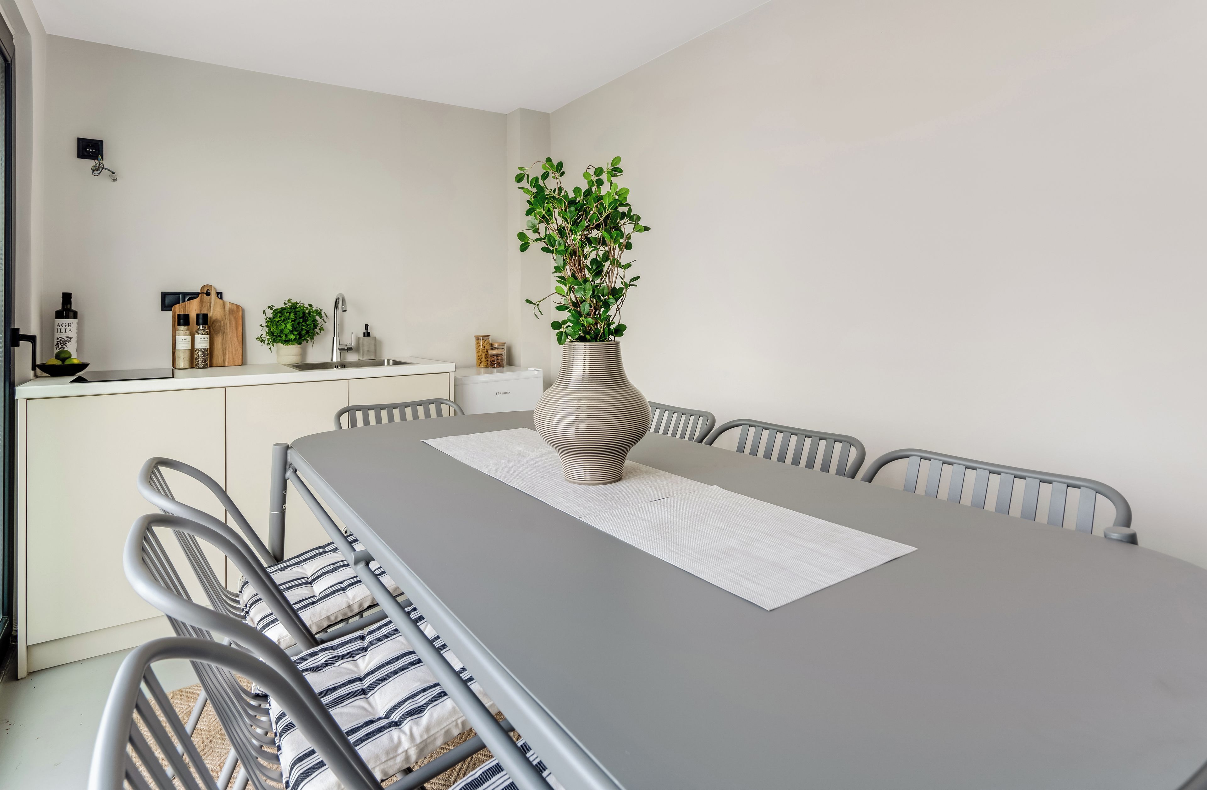 A modern dining area with a grey table, metal chairs, a vase with green plants, and a kitchen in the background.