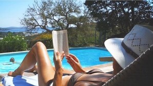 A person relaxing by a poolside with a book, wearing a hat, and a swimsuit. - Apartment-Standard-Ensuite with Shower-Sea View-T2 (Porto-Vecchio)