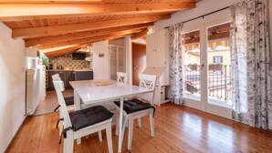 Um quarto aconchegante com teto de madeira, uma mesa de jantar com cadeiras, uma cozinha ao fundo e uma varanda com cortinas estampadas.