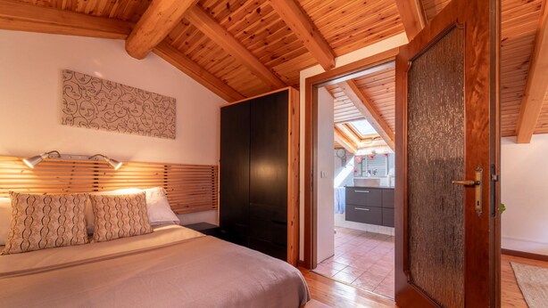 Quarto com teto de madeira, cama com almofadas estampadas, guarda-roupa preto e vista para uma cozinha através de uma porta aberta.