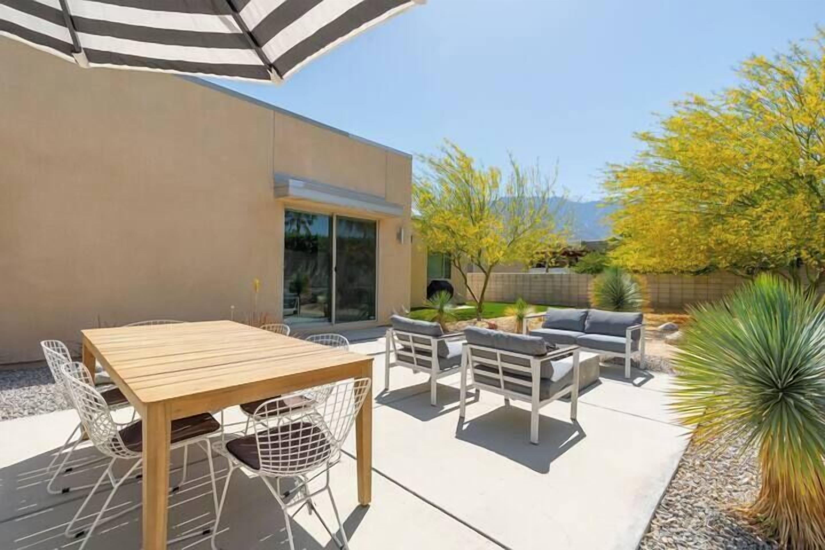 A patio with a wooden table and white chairs, a gray sofa, and a striped umbrella.