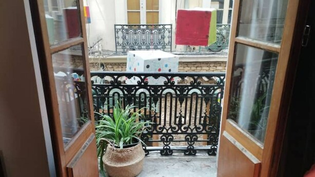 A view through a wooden-framed window with a wrought iron balcony railing, a potted plant, and a red object on the balcony. - Appartement en Plein Centre (Carpentras)