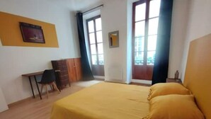 A room with a bed, a desk, and a chair. There are two windows with curtains. - Appartement en Plein Centre (Carpentras)