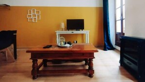 A living room with a wooden coffee table, a white TV stand with a flat-screen TV, a small white shelf, and a window with a blue curtain. - Appartement en Plein Centre (Carpentras)