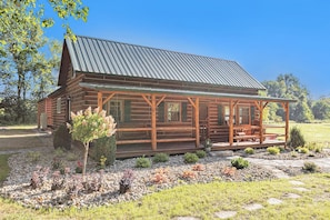 A log cabin with a porch and a metal roof, surrounded by a landscaped garden with various plants and a stone pathway.