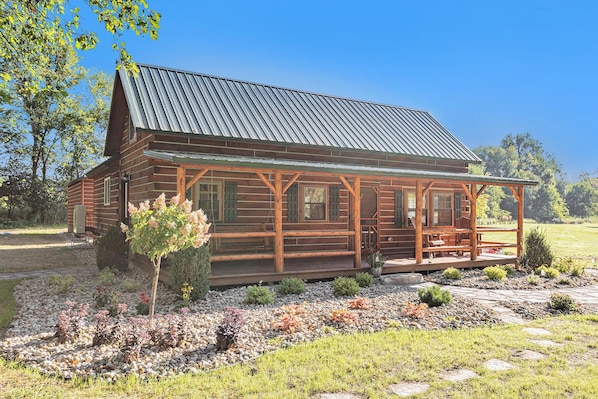 A log cabin with a porch, surrounded by a landscaped garden with various plants and a stone pathway.
