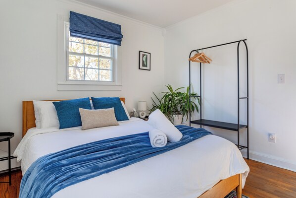 A bedroom with a large bed, a window with a blue valance, a bedside table, a hanging rack, and a potted plant.