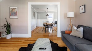 Una sala de estar con un sofá gris, una mesa de centro blanco y negro, piso de madera, un comedor con una mesa y sillas, y una planta en maceta.