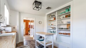 Una cocina con armarios blancos, encimera de madera, alfombra a rayas y una unidad de estanterías blancas con vajilla y utensilios de cocina.