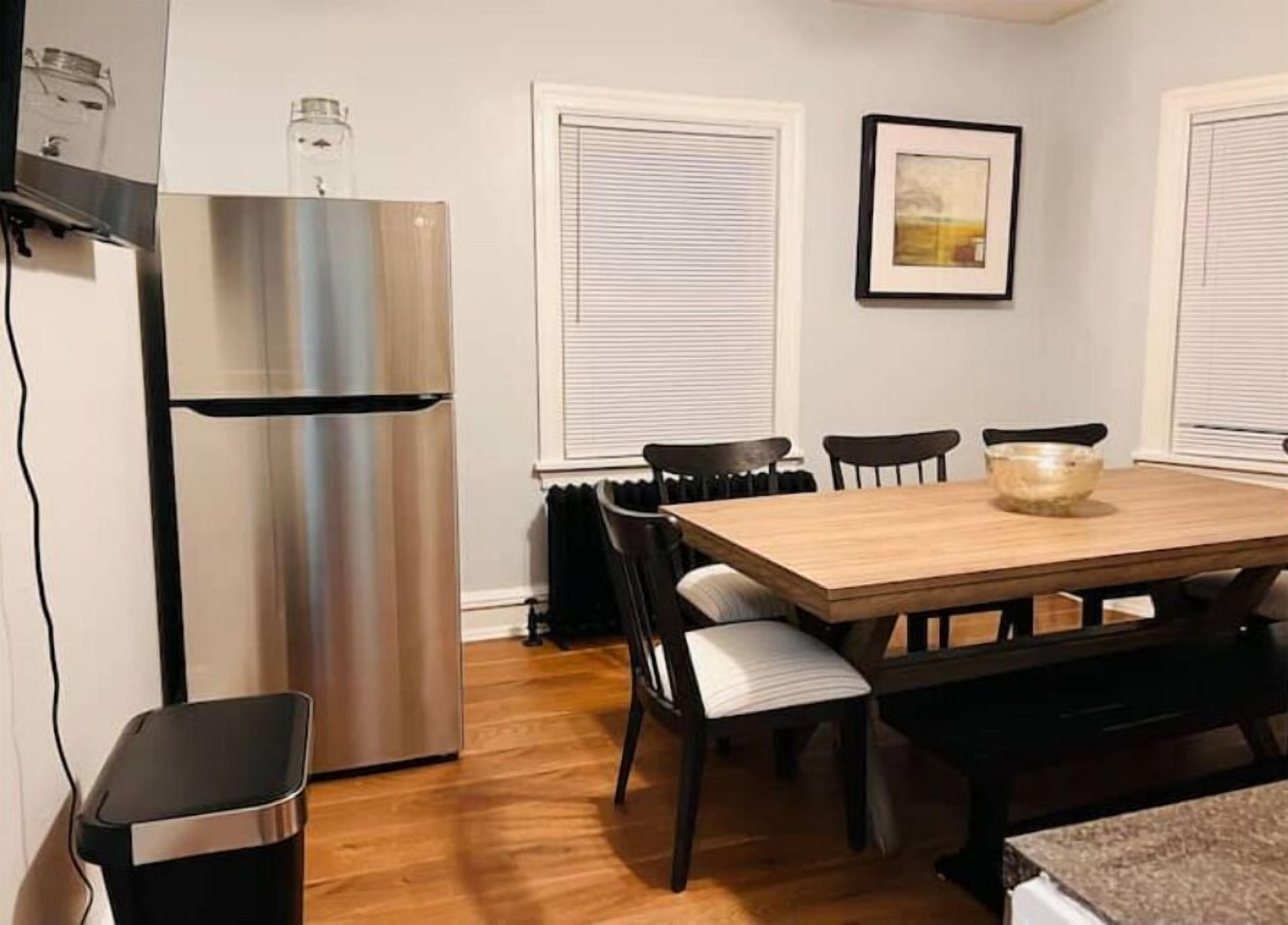 A compact kitchen with a stainless steel refrigerator, a wooden dining table with four chairs, and a black trash bin.