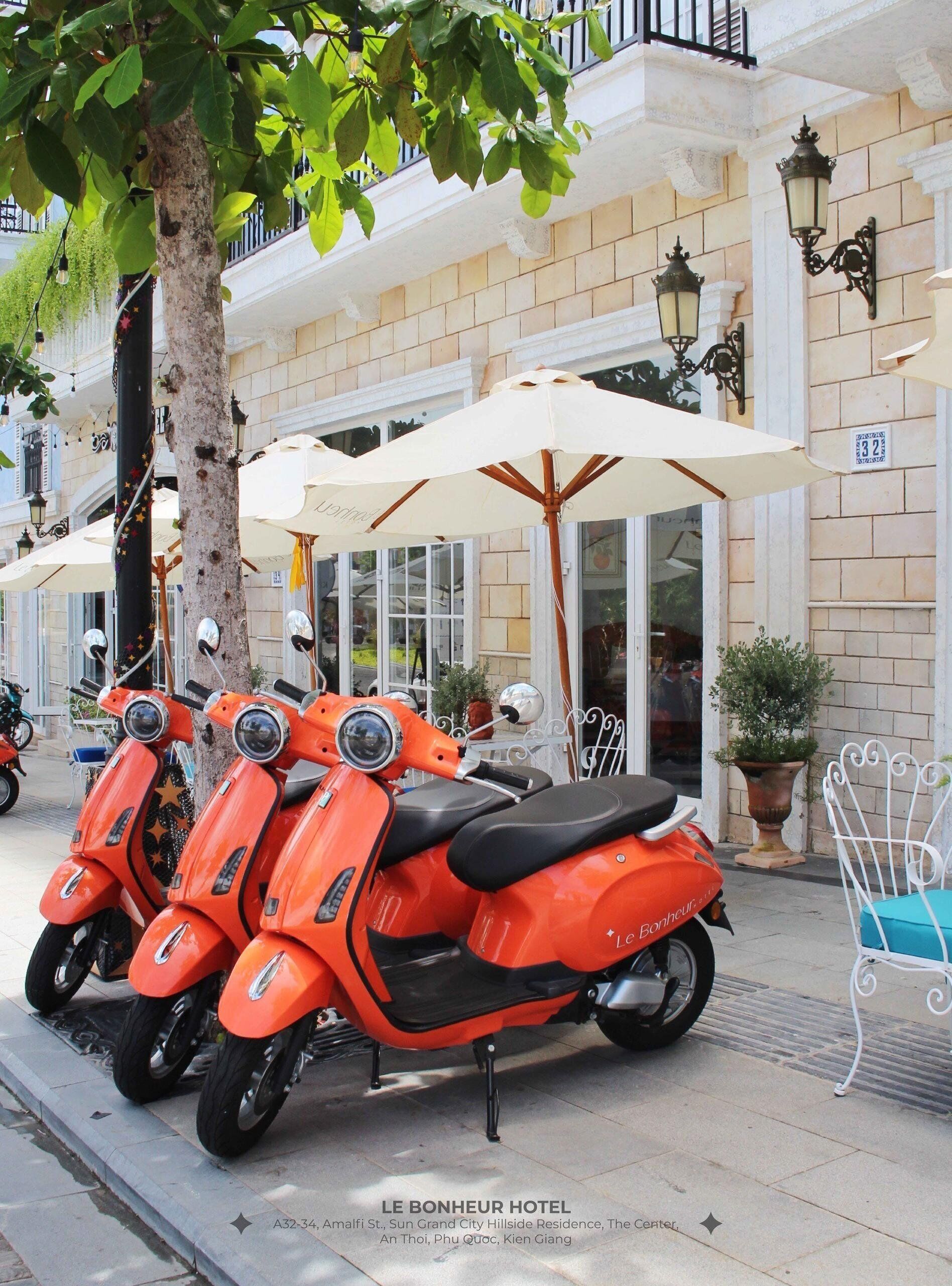 Uma fileira de scooters laranjas estacionadas em frente ao Hotel Le Bonheur, com guarda-sóis brancos e uma árvore por perto.