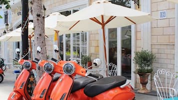 Uma fileira de scooters laranjas estacionadas em frente ao Hotel Le Bonheur, com guarda-sóis brancos e uma árvore por perto.