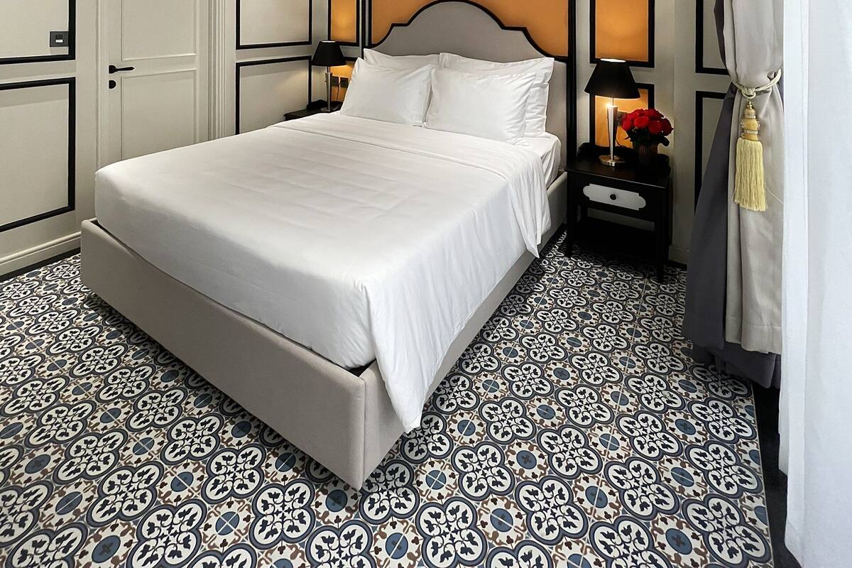 A neatly arranged bedroom with a large bed, a nightstand with a lamp, and patterned floor tiles.