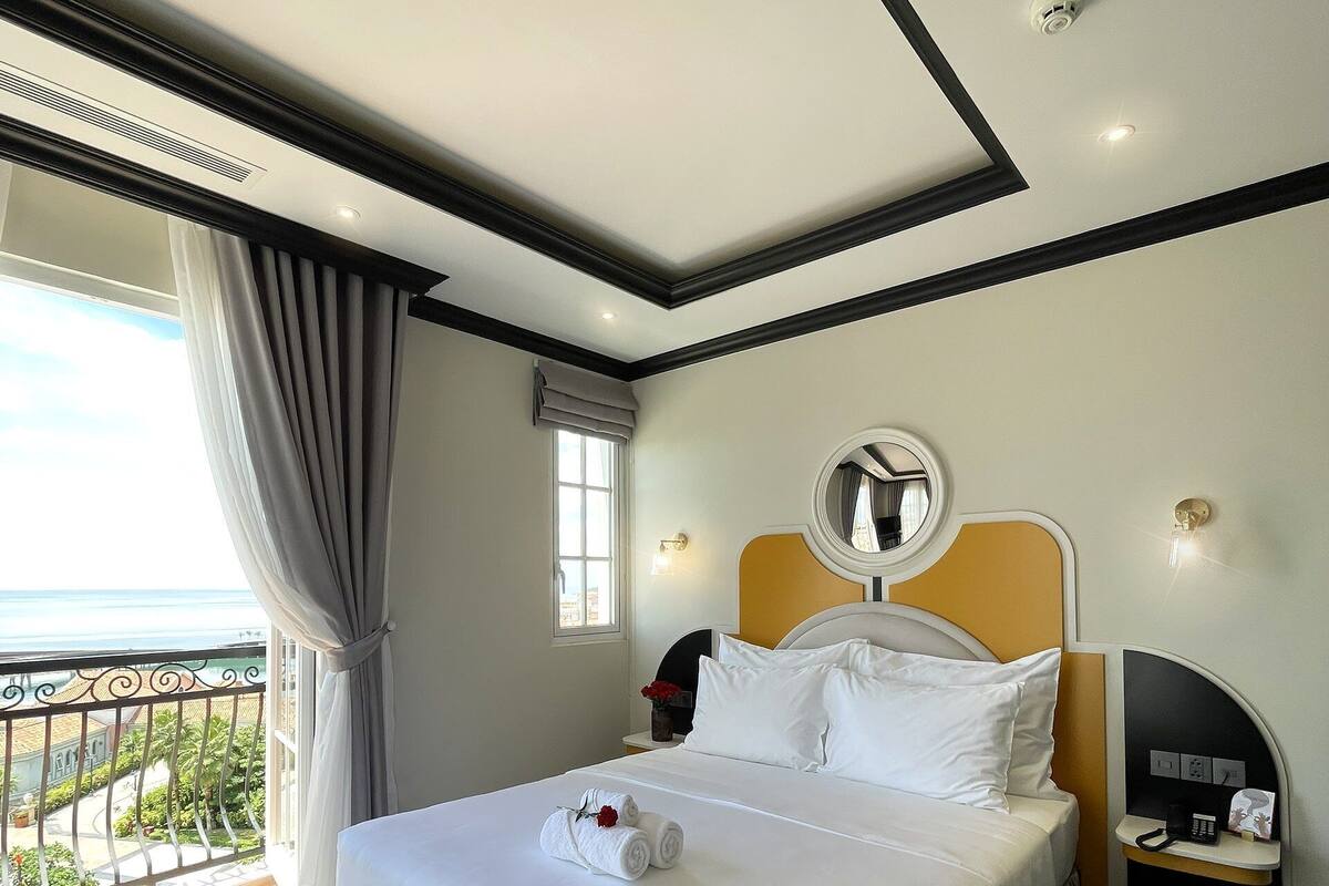A hotel room with a large bed, a bedside table with a phone and a lamp, a window with curtains, and a view of the outside.