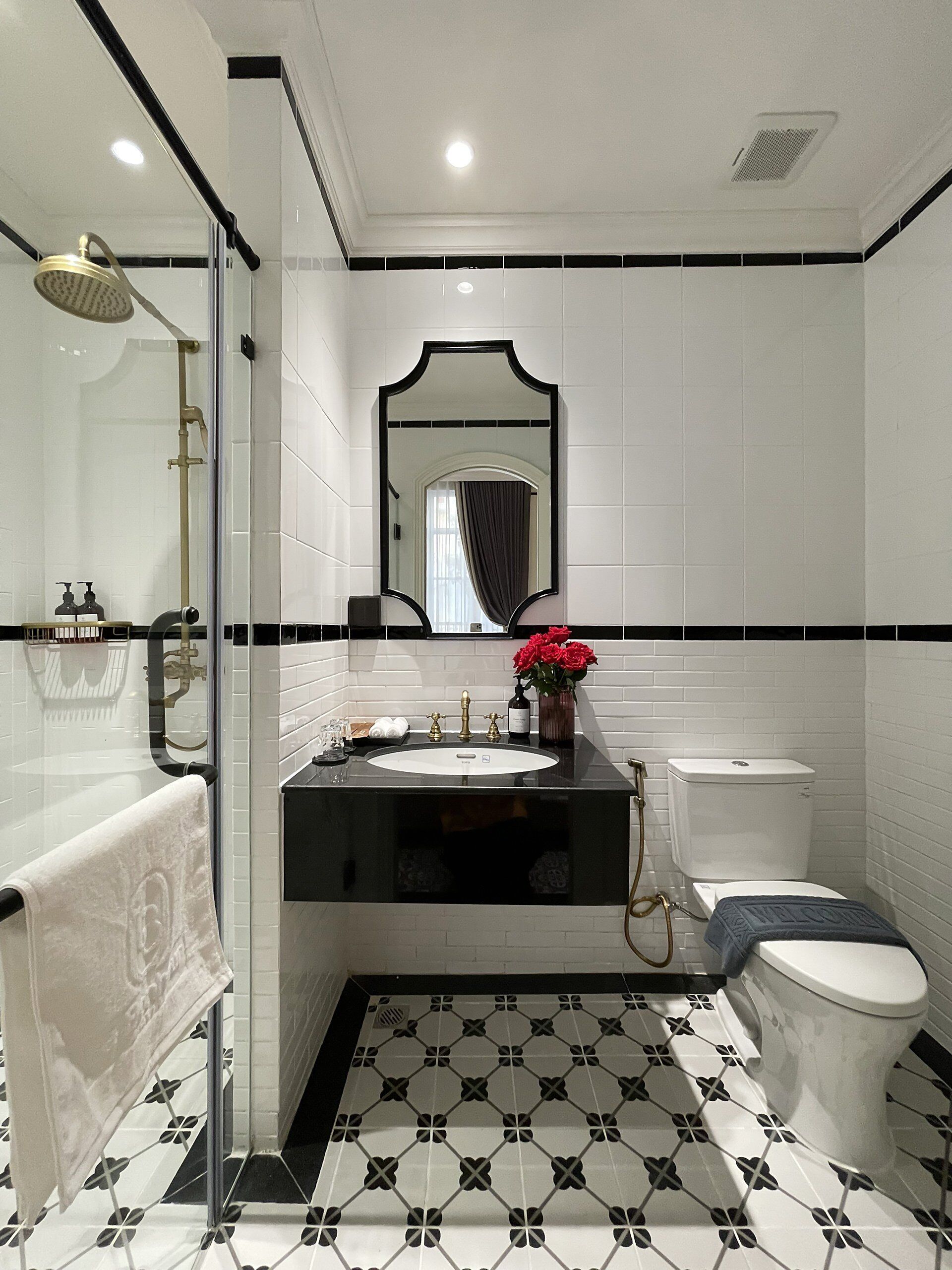 A modern bathroom with a black and white tiled floor, a white sink with gold fixtures, a glass-enclosed shower, and a toilet.