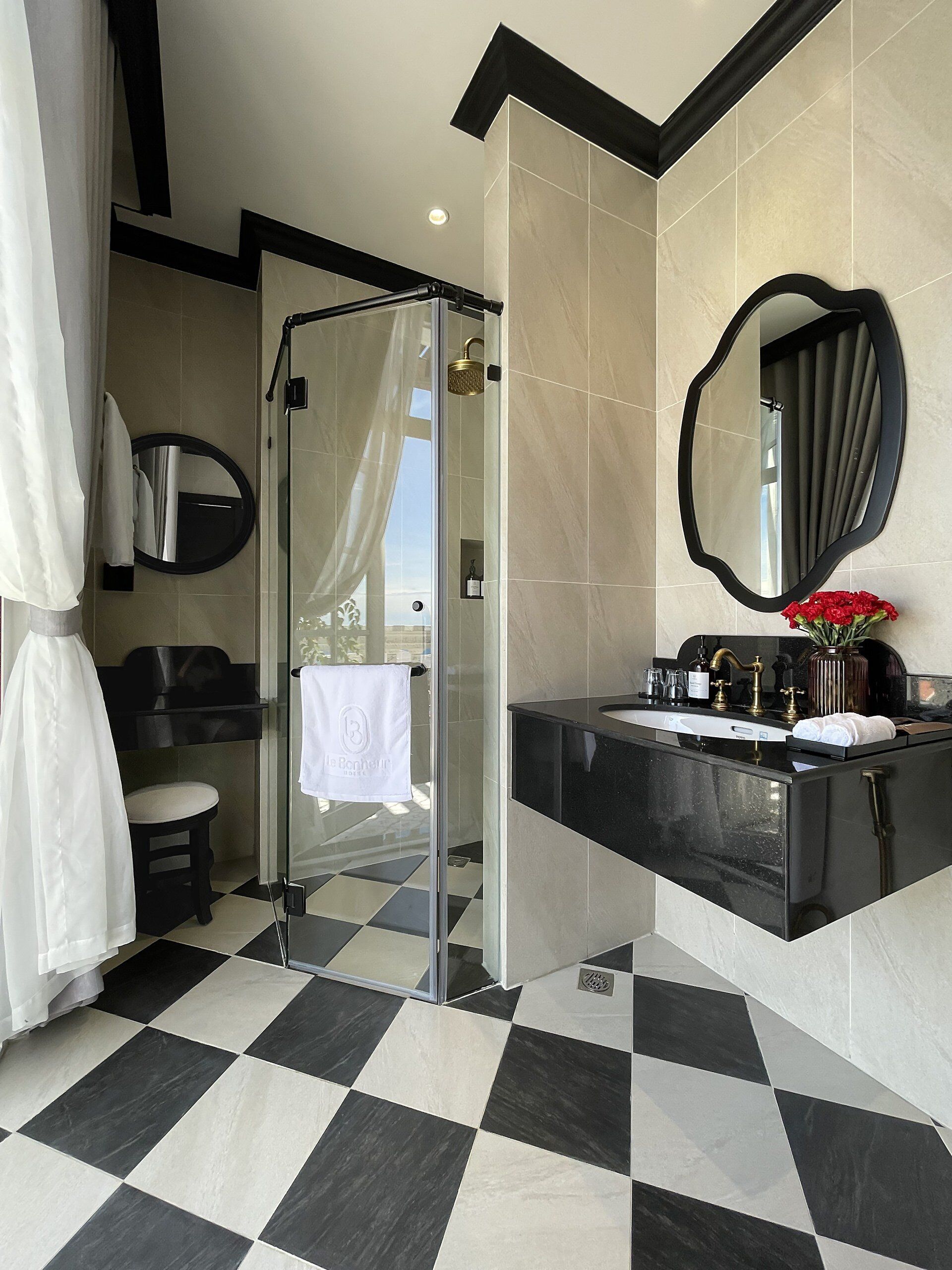 Un baño moderno con ducha de vidrio, piso a cuadros blanco y negro, espejo redondo y tocador con encimera negra.