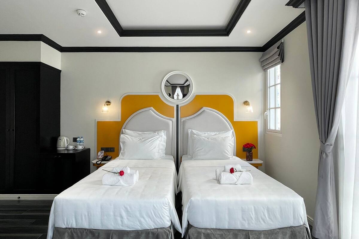 A hotel room with two beds, a round mirror above the beds, a small table with a kettle and cups, and a window with curtains.