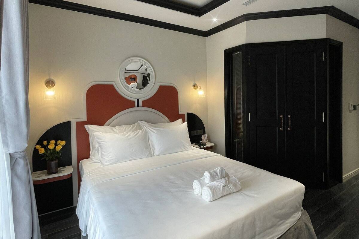 A modern hotel room with a large bed, a bedside table with a lamp, a wardrobe, and a mirror on the wall.
