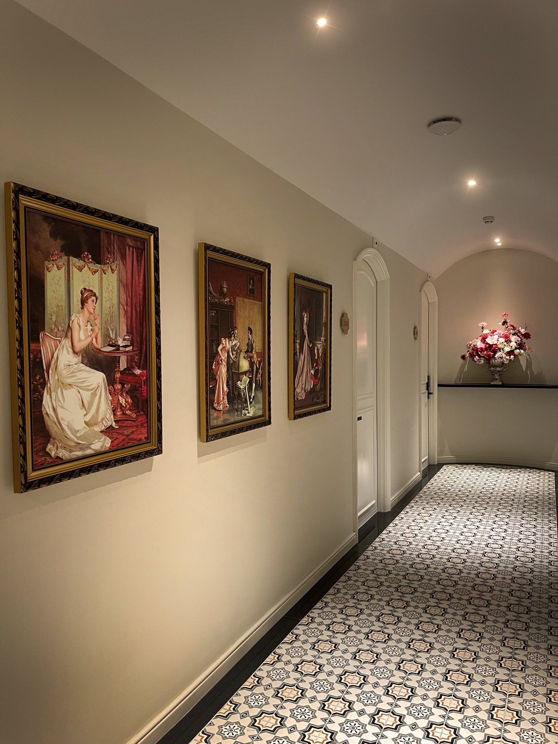 A hallway with three framed paintings on the wall, a white door, and a floral arrangement on a shelf.