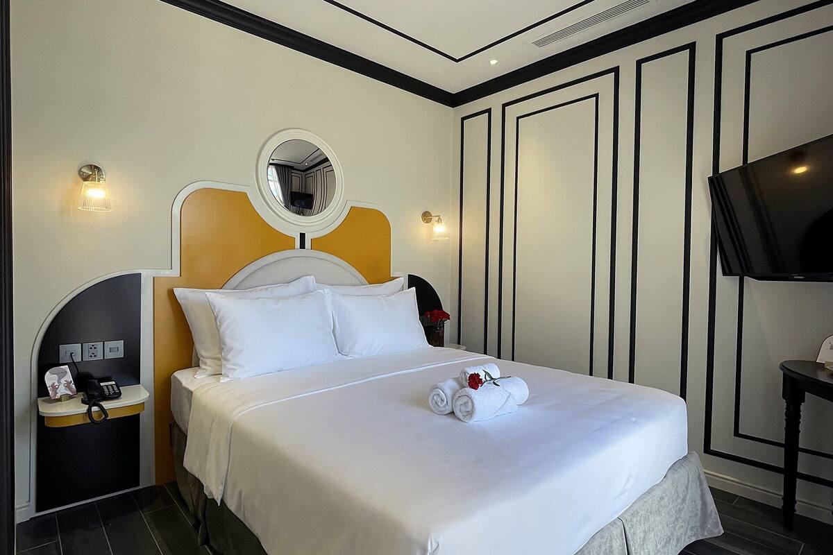 A modern hotel room with a large bed, a bedside table with a phone, a mirror, and a flat-screen TV.