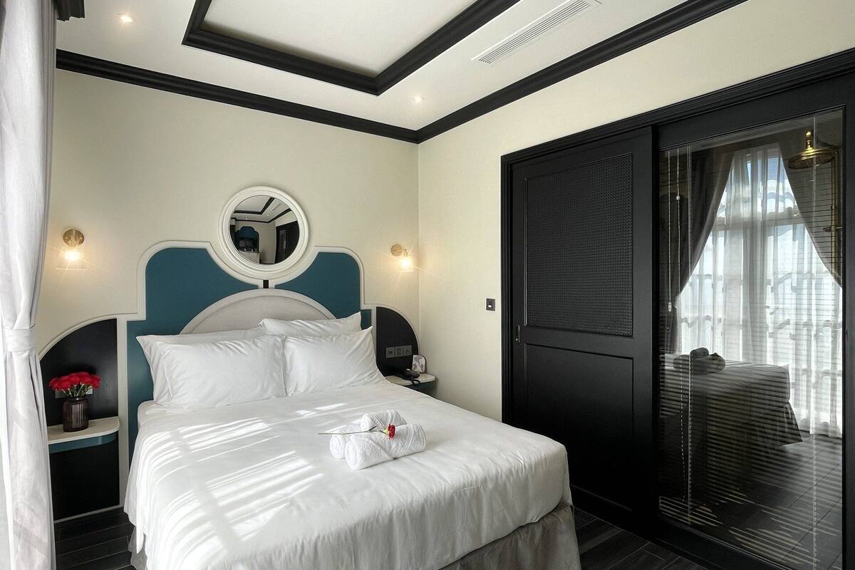 A modern hotel room with a large bed, a bedside table with a lamp, a mirror, and a sliding glass door to a balcony.