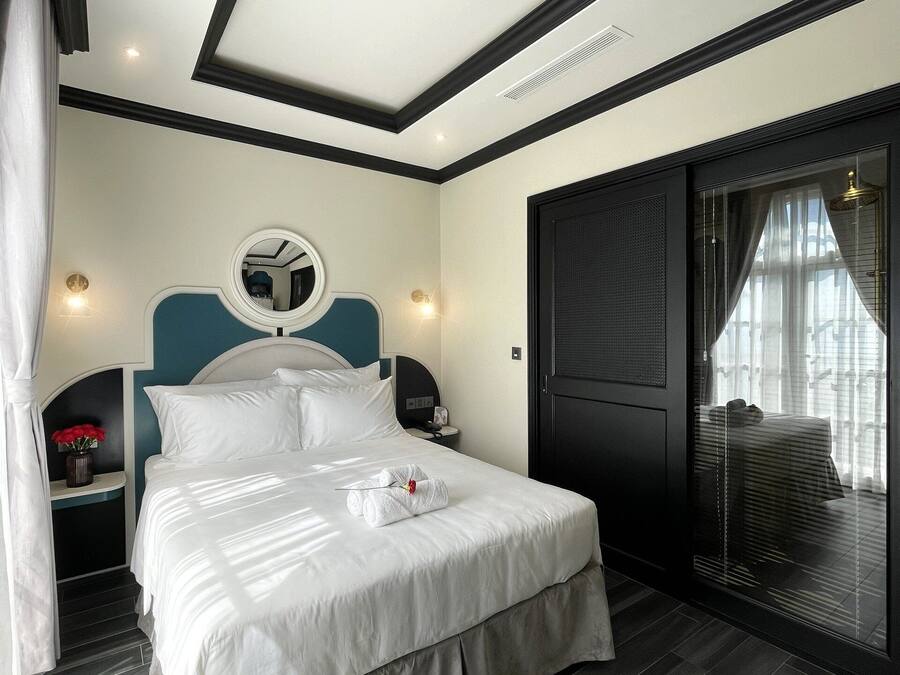 A modern hotel room with a large bed, a bedside table with a lamp, a mirror, and a sliding glass door to a balcony.
