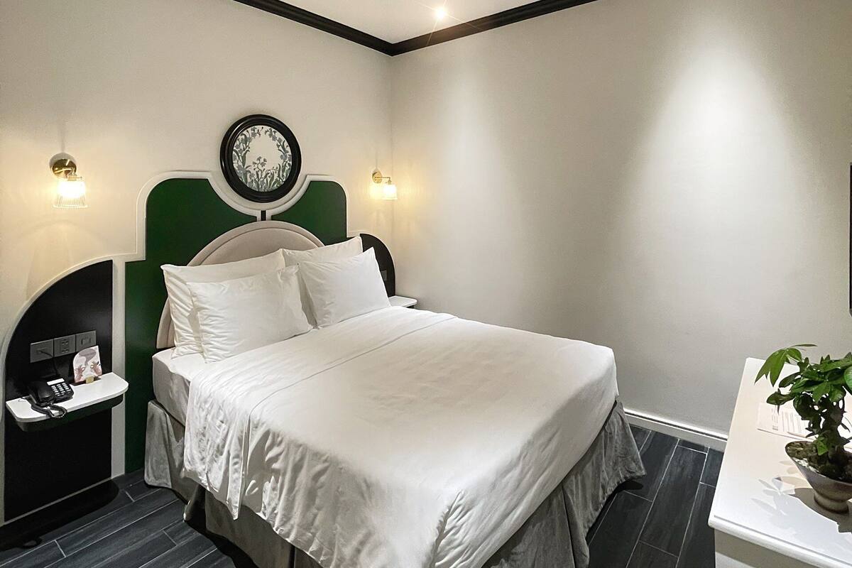 A modern hotel room with a large bed, a bedside table with a lamp, a wall clock, and a small plant on the nightstand.