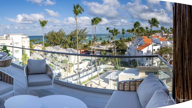 A balcony with wicker furniture, a glass railing, and a view of the ocean and palm trees. - Luxurious Chic Penthouse Navio Beach (Punta Cana)