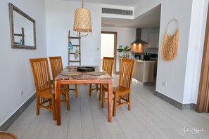Uma área de refeições com mesa e cadeiras de madeira, uma luminária pendente de fibra trançada e uma cozinha ao fundo.