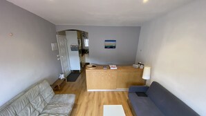 Een moderne zithoek met een bank, een klein tafeltje en een blauwe divan. Er is een keuken te zien op de achtergrond met een welkomstbord.