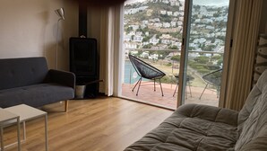 Een moderne woonkamer met een bank, een fauteuil en een tv-meubel. Een groot raam biedt uitzicht op een heuvel met gebouwen.