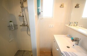 Baño con ducha, lavabo y espejo. Hay artículos de aseo en el lavabo y un perchero para toallas.