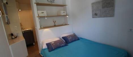 A bedroom with a bed, a nightstand, a shelf with decorative items, and a wall-mounted light fixture.