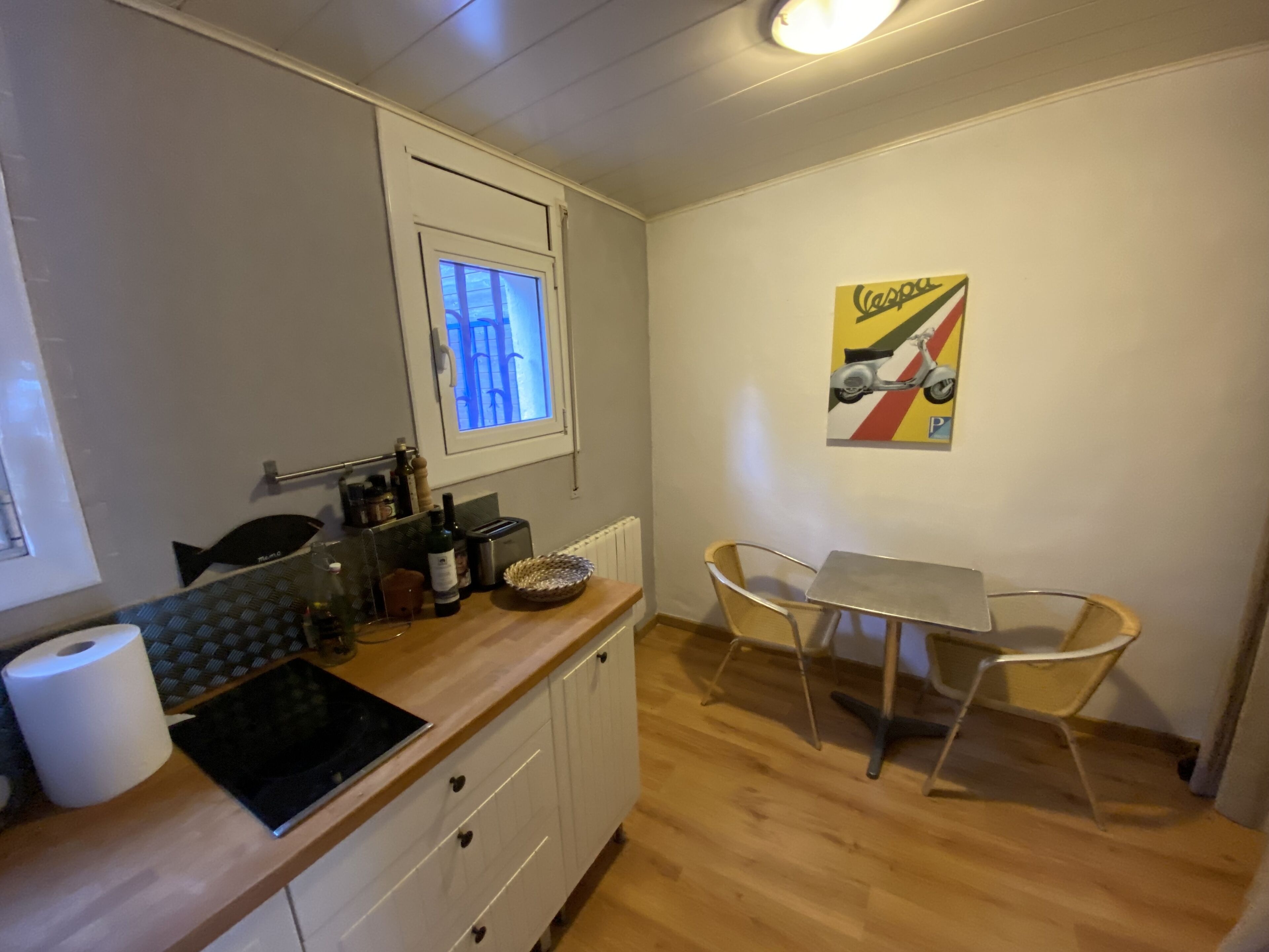 Une cuisine de taille réduite avec une fenêtre, un coin repas comprenant deux chaises, une table et un tableau accroché au mur.