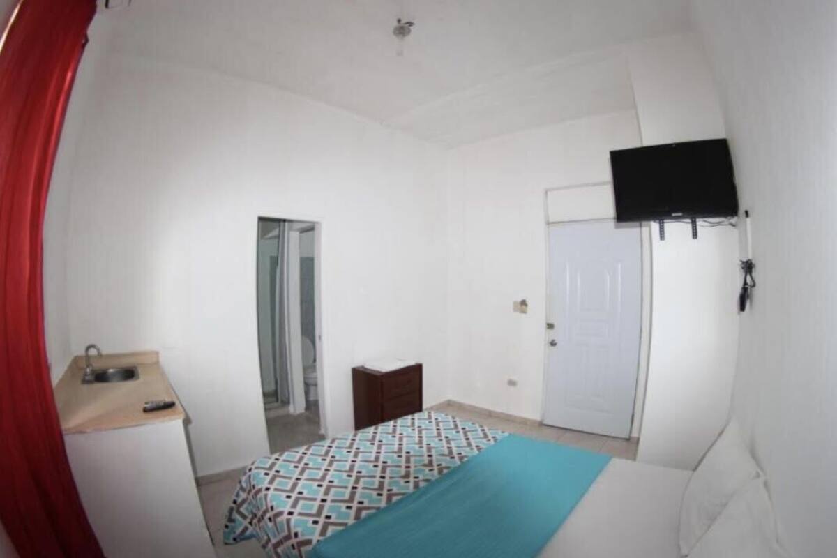 Une petite pièce propre, avec un lit, un bureau doté d’un lavabo, un miroir, une porte et un téléviseur fixé au mur.