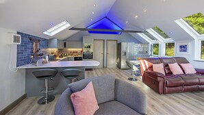 Una cocina moderna con una isla central, taburetes de bar y una zona de estar con un sofá de cuero y un sillón gris.