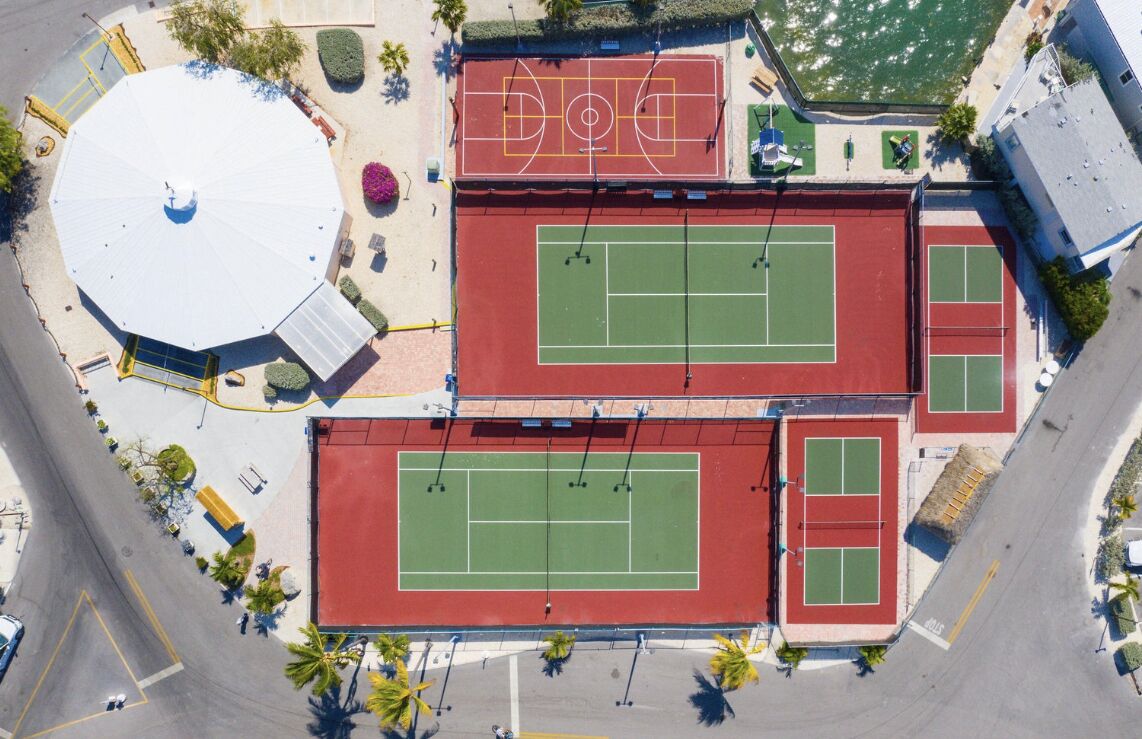Vue aérienne d’un complexe sportif comprenant plusieurs courts de tennis, un terrain de basketball et une zone couverte équipée de panneaux solaires.