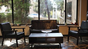 Una habitación con suelo de piedra, un sofá, dos sillones, una mesita con un objeto de vidrio y amplias ventanas que dan a árboles y coches aparcados.