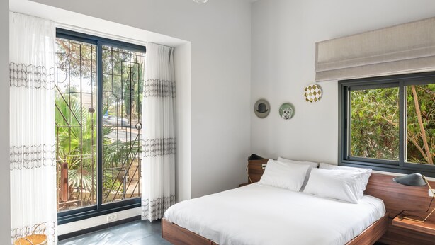Un dormitorio con un ventanal grande, una cama con ropa de cama blanca, una mesita de noche con lámpara y vistas al exterior con vegetación.