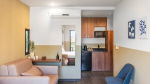 Una habitación de hotel moderna con sofá, una silla, una mesita con velas, un espejo y una kitchenette con microondas y refrigerador.