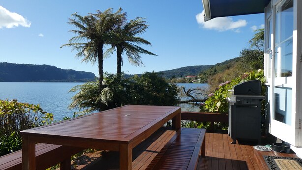 Een houten terras met een tafel en banken met uitzicht op een meer, omringd door weelderige begroeiing en een vergezicht op verre heuvels.