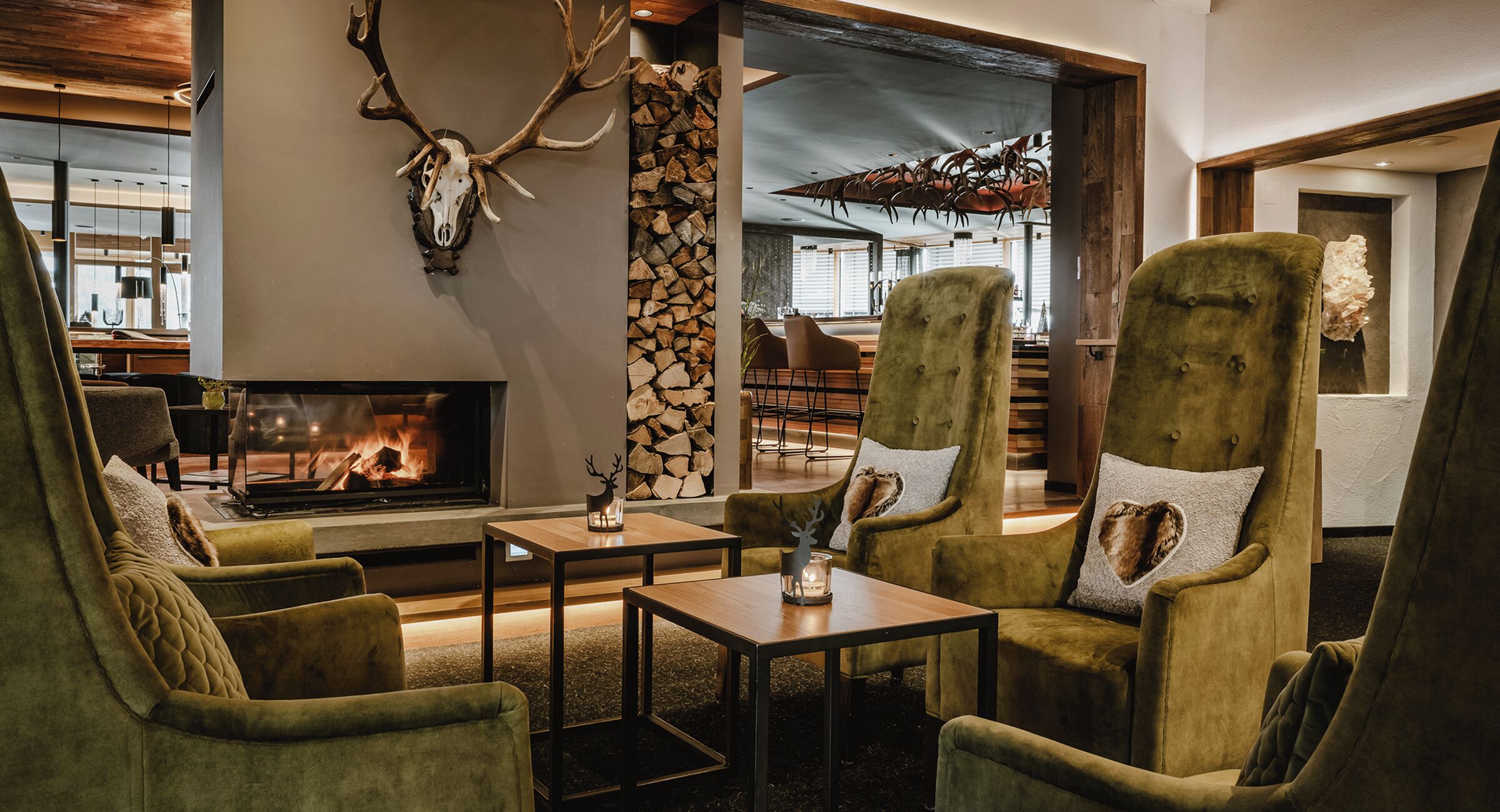 A cozy lounge area with plush green armchairs, a fireplace, and a stone wall with a deer skull.