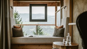 Un coin salon confortable avec vue sur un paysage montagneux, une table en bois avec une bouteille et un verre, et une fenêtre à cadre noir.