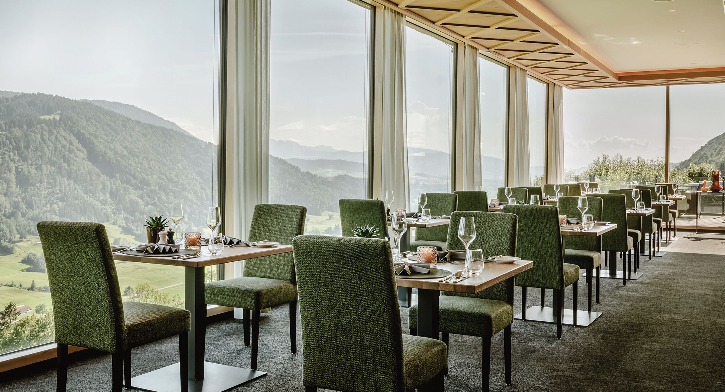 A restaurant with a mountain view, green upholstered chairs, and tables set for dining.