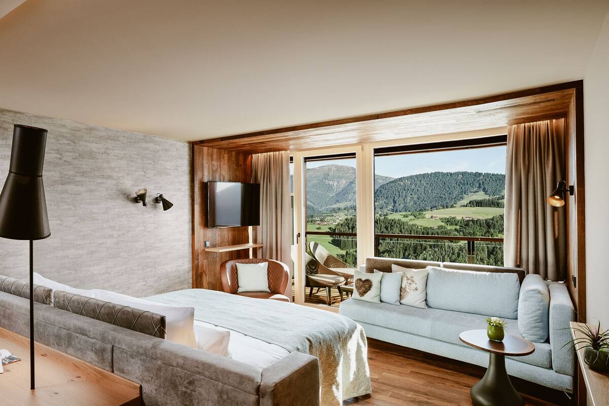 Habitació d'hotel moderna amb un llit gran, un sofà, zona de menjador i un balcó amb vista panoràmica a les muntanyes.