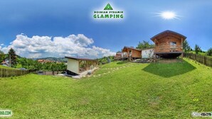 A glamping site with wooden cabins on a grassy hill, a clear sky, and a logo for Bosnia Pyramid Glamping.