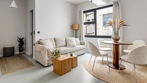 Ein modernes Wohnzimmer mit einer Couch, einem Couchtisch und Sesseln, einem runden Holztisch und einem großen Fenster mit Vorhängen.