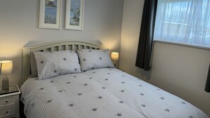 A bedroom with a bed, two framed pictures on the wall, a bedside lamp, and a window with curtains. - M & T Chalets , 2 bed chalet Hemsby. sleeps 6 (great yarmouth)